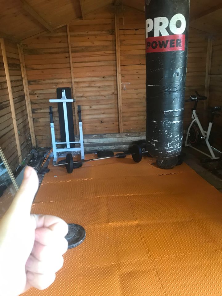 a room with orange mats on the floor, a punch bag in the front right fore ground, weightlifting equipment in the back left and a thumbs up in the left fore ground